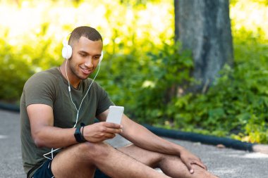 Rahat siyah adam koşu pistinde oturuyor ve parkta akıllı telefondan müzik dinliyor.