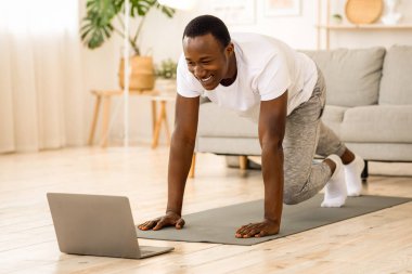 Oturma odasında egzersiz yapan neşeli siyahi adam dizüstü bilgisayara bakıyor. Neşeli Afrikalı Amerikalı genç adam çevrimiçi fitness dersleri alıyor, vücudunu dizüstü bilgisayarın önünde geriyor, uzayı kopyalıyor.