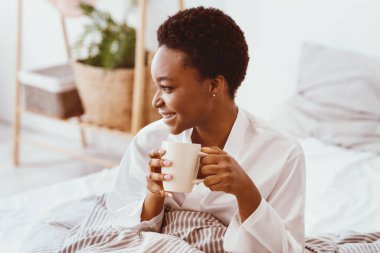 Kahvaltıda kahve, favori latte, zindelik ve sabahları enerji. Neşeli, genç, Afro-Amerikan bayan, yatak odasının içinde, sıcak bir bardak içecekle yatakta oturuyor.