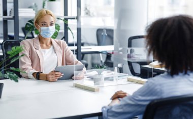 Patronla konuşmak ya da koronavirüs salgını sırasında röportaj yapmak. Koruyucu maskeli, tabletli genç bir kadın modern ofis içi koruyucu cam aracılığıyla Afro-Amerikan bayanla iletişim kuruyor.