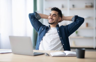 Mutlu genç Arap adam işini seviyor, çalışma masasında oturuyor, arkasına yaslanıyor, fotokopi odasına bakıyor ve gülümsüyor, ofis içi. Milenyum için iş fırsatları, kariyer kavramı