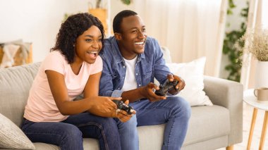 Neşeli Afro-Amerikan aile babası karı-koca evde video oyunları oynuyor, fotokopi odası. Genç ve mutlu siyah erkek ve kadın oyuncular oturma odasında kanepede oturuyor ve ellerinde joysticklerle rekabet halindeler.