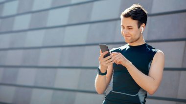 Müzikle modern çalışma. Gülümseyen genç adam telefon, fitness takip cihazı ve kablosuz kulaklıkla gri duvar arka planında çalışmaya hazırlanıyor.