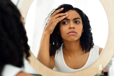 İnce çizgiler ve kırışıklıklar. Endişeli siyahi kadın aynaya bakıyor. Yüzündeki yaşlanma işaretlerini kontrol ediyor, seçici odaklanıyor.