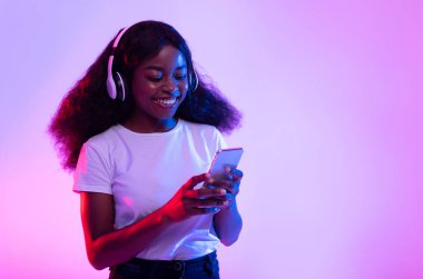 Kablosuz kulaklıklı, neon ışıkta akıllı telefon kullanan neşeli, bin yıllık siyahi kadın, kopyalama alanı. Güzel Afro-Amerikan kadın yeni mobil müzik uygulamasını kontrol ediyor.