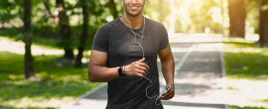 Cep telefonuyla koşan, müzik dinleyen ve güneşli bir sabahta parkta koşmanın keyfini çıkaran mutlu bir koşucu. Panorama