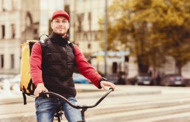 Bisikletli kurye elinde çantayla şehre yiyecek getiriyor, kameraya gülümsüyor. Metin için Boş Alan
