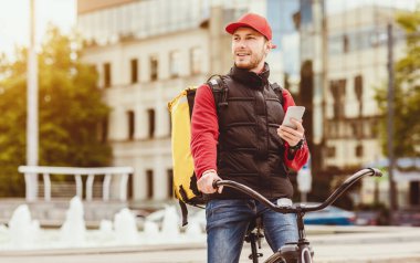 Teslimat Uygulaması Konsepti. Kurye adam cep telefonu kullanıyor yiyecek teslimatı yapıyor bisikletle şehir dışına çıkıyor. Boşluğu Kopyala