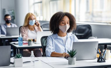 Coronavirüs salgınında sosyal mesafeler. Koruyucu maskeli güzel Afro-Amerikan bayan dizüstü bilgisayarda yazıyor, şirket destek merkezinde çalışıyor diğer çalışanlarla birlikte.