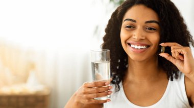 Sağlık Konsepti. Gülümseyen Afrikalı Kadın Multivitamin Kapsülü ve bir bardak su tutuyor, Sağlıklı yaşam tarzının tadını çıkarıyor, Kopya Boşluğu