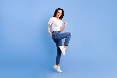 Neşeli genç bir kadının komik portresi. Turkuaz arka planda izole edilmiş yumruklarıyla zıplıyor, bir bacağını kaldırıyor.