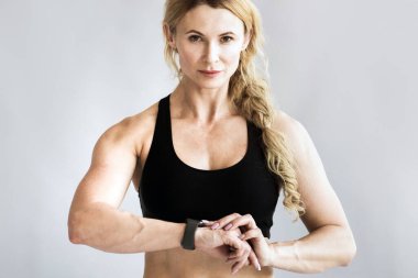 Covid-19 tecridi boyunca formda kalın ve orta yaşlarda evdeki tavan arasında modern antrenman yapın. Spor giyimli, zayıf, kaslı, yetişkin bir kadın spor giyim sektöründe nabzını kontrol ediyor ve kameraya bakıyor.