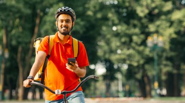 Hızlı servis. Parkta arkadaş canlısı kurye, kasklı, bisikletli, akıllı telefon tutan ve müşteri adresi arayan, boş alan arayan.