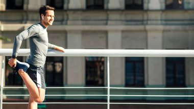 Ciddi genç atlet dışarıda esneme hareketleri yapıyor. Kablosuz kulaklıklı adam, sabah koşmadan önce şehrin arka planında, profilinde, boş alanda ısınıyor.