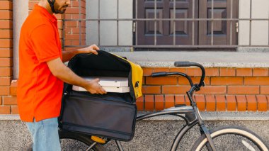 Eve ve ofise teslimat. Kasklı genç teslimatçı bisikletli büyük sarı sırt çantasından pizzayı alıyor, ön kapının yanında, yan manzara, serbest alan.