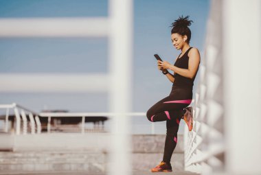 Açık havada müzik eşliğinde sabah antrenmanı. Spor üniformalı Afrikalı Amerikalı bir kız, kablosuz kulaklıkla duvara yaslanıyor ve telefona bakıyor.