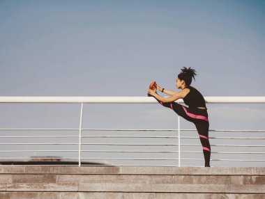 Spor tarzı, antrenman ve egzersiz. Spor üniformalı, zayıf Afrikalı Amerikalı kız stadyumda bacaklarını esnetiyor, mavi gökyüzü arka planı, boş alan.