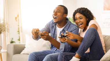 Ev eğlencesi. Keyifli Afro-Amerikalı erkek ve kadın ellerinde joysticklerle oturma odasında oturmuş video oyunları oynuyor ve gülüyorlar. Kilitleme sırasında evde birlikte eğleniyorlar.