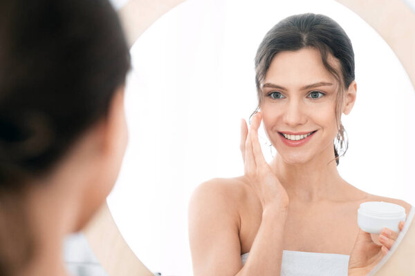 Good looking young lady using face cream at home, looking at mirror and smiling, shot from behind. Smiling woman with ponytail holding cream jar and applying beauty product on her cheeks