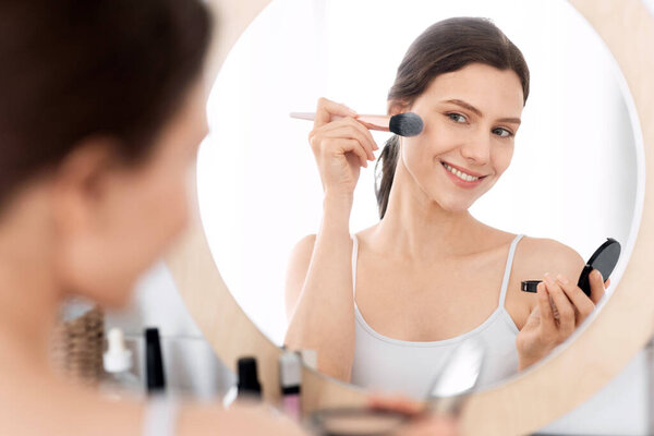 Happy pretty young woman applying blush on her face, sitting in front of mirror, home interior, copy space. Smiling attractive young lady doing makeup before going out, shot over shoulder in bedroom