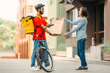 Bisikletli kurye müşteriye isim çantası getirmemiş. Gülümseyen kız, kasklı, sırt çantalı, yan manzaralı, boş alan olan genç adamdan kağıt torba alır.