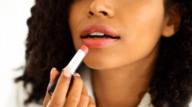 Günlük makyaj. Bornozlu tanınmayan siyahi kadın pembe ruj sürüyor, evde dudaklarıyla ilgileniyor, kesilmiş görüntü, yakın plan.