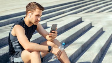 Enerji, zindelik ve aksiyon. Kablosuz kulaklıklı ve fitness izleyicili bir sporcu elinde bir şişe su ve akıllı telefon tutuyor, merdivenlerde oturuyor, boş yer var.