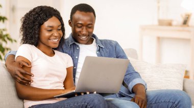 Gülümseyen siyah çift evdeki kanepede dinlenirken dizüstü bilgisayar kullanıyor, birlikte komik içerik izliyorlar, uzayı kopyalıyorlar.