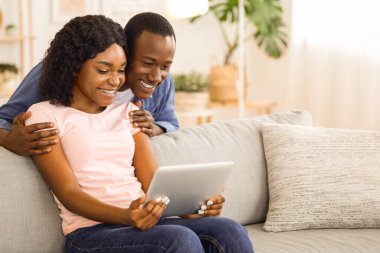 Heyecanlı siyah çift dijital tablet ekrana bakıyor, yeni uygulama kullanıyor, oturma odasında kanepede oturuyor, boş bir alan. Memnuniyetle Afro-Amerikan erkek ve kadın evde birlikte eğleniyorlar.
