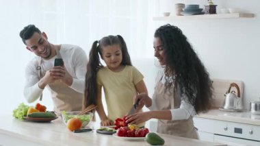 Orta Doğulu bir aile, taze sebzelerle bir yemek hazırlamak için modern bir mutfakta toplanıyor. Anne, kızının malzemelerini doğramasına yardım ederken baba telefonuyla bir an yakalıyor..