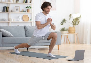 Neşeli sporcu adam evde kilitli kalırken egzersiz yapıyor, dizüstü bilgisayar kullanıyor, boş alan kullanıyor. Oturma odasında atletik erkek eğitimi, spor dersleri veya yoga dersleri, sağlıklı yaşam tarzı konsepti.