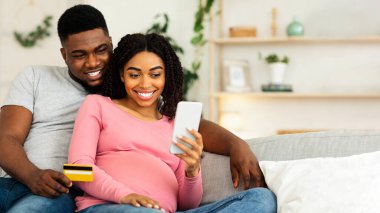 Mutlu hamile çift online alışveriş için mobil uygulama kullanıyor, evde oturuyor, kredi kartı kullanıyor, boş alan kullanıyor.