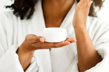 Siyahi kadın elinde organik cilt kremi kavanozuyla duştan sonra yüzüne nemlendirici sürüyor bornoz giyiyor ve seçici bir odak noktası olan kesilmiş bir görüntü sergiliyor.