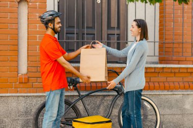 Güvenli bir teslimat. Kasklı gülümseyen kurye, bisikletli büyük sarı sırt çantalı, müşteriye ön kapıda, dışarıda, yan tarafta, boş alanda kağıt torba veriyor.