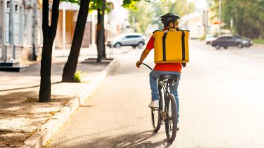 Bisikletli kurye, paket, bisiklet, teslimat servisi. Üniformalı, koruyucu kasklı, yazın şehirde yolda sarı termo çanta sürücüleri olan ve müşteri adresini, boş alanı, arka tarafı arayan genç bir adam.