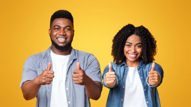 Genç bir adam ve kadın yan yana durur, gülümser ve başparmak kaldırırlar. Günlük kıyafetleri ve parlak ifadeleri canlı sarı bir ortamda pozitif ve canlandırıcı bir atmosferi vurguluyor..