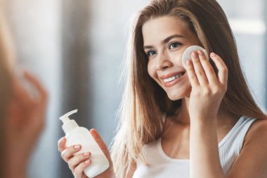 Güzel genç bir kadın banyoda aynanın önünde makyaj losyonu ya da makyaj losyonu kullanıyor.