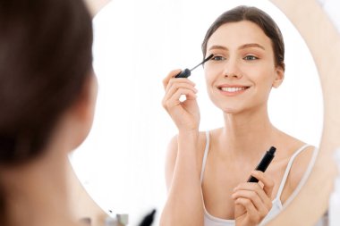 Gülümseyen genç bir kadın makyaj yapıyor, aynaya bakıyor, maskara kullanıyor, ev dekorasyonu yapıyor.