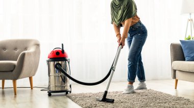 Genç bir kadın aktif olarak evinin oturma odasını süpürüyor. Günlük kıyafetler giyiyor ve halıyı temizlemeye odaklanıyor. Temiz ve düzenli bir yer yaratıyor..