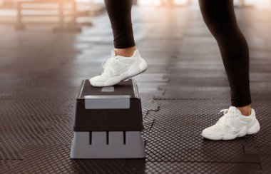 Spor salonundaki yükseltilmiş bir platformda spor ayakkabıları ve siyah taytlar giyerek egzersiz yapan bir insan. Çevre iyi aydınlatılmış ve fitness eğitimi için tasarlanmıştır..