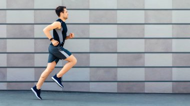 Sağlık için koşuyorum. Spor kıyafetli genç adam, spor takip cihazı gri duvarda çalışıyor.