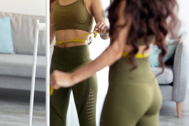 Antrenman kıyafeti giymiş genç bir Hintli kadın aynanın önünde duruyor ve kemerini mezurayla ölçüyor. Sağlıklı yaşam ve spor yapmaya olan bağlılığını yansıtıyor..