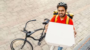 Sipariş teslimi. Kasklı, sakallı, gülümseyen genç adam, gözetleme deliğine pizza kutusu gösteriyor, sokakta bisikletin yanında, boş yer var.
