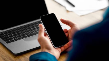 Genç bir Orta Doğulu girişimci, çağdaş bir ofiste masa başında otururken boş ekranla akıllı telefonuna odaklanıyor. Şirket ortamında iş ve iletişimi dengeliyor..