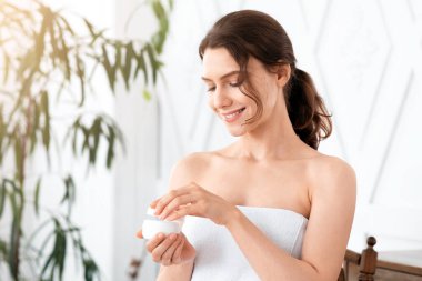 Havluya sarılmış gülümseyen kadın vücut ya da yüz kremi kullanıyor, yatak odasının içinde. Güzel bir bayan sabah duştan sonra yüzü ya da vücudu için nemlendirici kullanıyor. Elinde krema kavanozu, kopyalama alanı var.