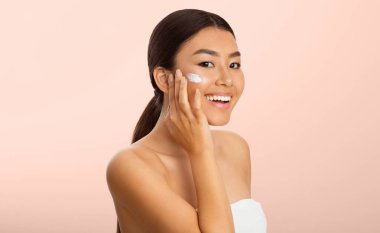 Güzellik Konsepti. Yüze Nemlendirici Uygulayan Asyalı Kadın, Pembe Stüdyo Arkaplanı