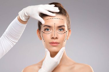 Estetik ameliyat kavramı. Genç bir kadın kozmetoloji kliniğinde tedavi görüyor, görünüşünü değiştiriyor. Plastik cerrah güzel kadın yüzüne izler bırakıyor, gri stüdyo arka planı, kopyalama alanı.