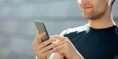 Antrenman için favori parça ve harika ruh hali. Spor kıyafetli, kablosuz kulaklıklı, gülümseyen adam akıllı telefona bakıyor, kapatıyor, boş alan, güneş parlıyor.