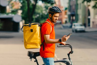 Modern mobil uygulamalar. Güvenlik kasklı, büyük sarı çantalı, bisikletli genç adam akıllı telefona, açık havaya, profile, kopyalama alanına bakıyor.
