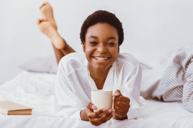 Güne olumlu bir başlangıç ve kahve. Gülümseyen Afrikalı Amerikalı genç kadın bornozla yatakta uzanıyor ve elinde fincan tutuyor, yakın plan, serbest alan.